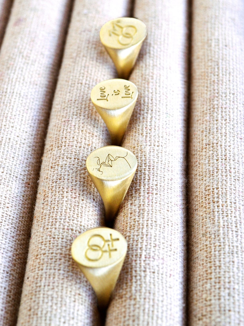 Puede incluir: Cuatro anillos de sello de oro con diferentes grabados. Los anillos se muestran en una fila sobre un fondo de tela beige. Los grabados incluyen las palabras "Love is Love", un par de corazones entrelazados, una imagen estilizada de dos personas bes&aacute;ndose y el s&iacute;mbolo femenino y masculino.