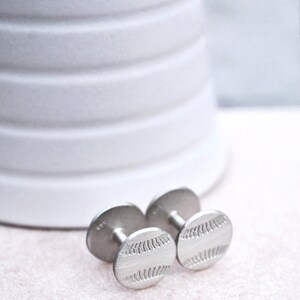 May include: A pair of silver cufflinks with a baseball design. The cufflinks are round and have a textured surface.