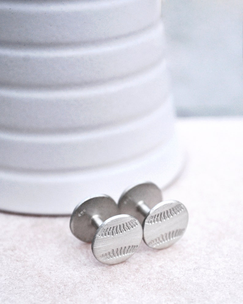 May include: A pair of silver cufflinks with a baseball design. The cufflinks are round and have a textured surface.