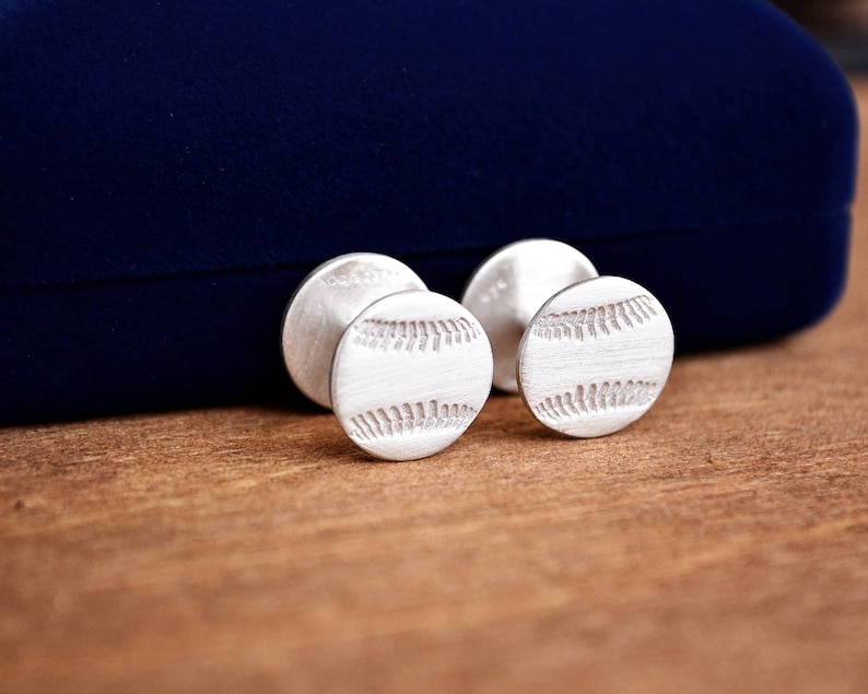 May include: A pair of silver cufflinks with a baseball design. The cufflinks are on a blue velvet background.