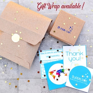 K&ouml;nnte beinhalten: Eine braune Papiert&uuml;te mit einem goldenen Sternmuster und einem goldenen runden Aufkleber. Die T&uuml;te liegt auf einer grauen Oberfl&auml;che mit goldenem Sternkonfetti. Ein kleiner brauner Karton mit dem Text "Bubble Box" ist ebenfalls sichtbar. Der Karton liegt auf einer blauen Karte mit einer Weltkarte und dem Text "Wie man f&uuml;r seinen Schmuck sorgt". Eine wei&szlig;e Karte mit dem Text "Danke!" und eine blaue Karte mit dem Text "Bubble Box" sind ebenfalls sichtbar.