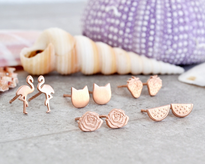 May include: A set of rose gold stud earrings featuring various designs, including flamingos, cats, strawberries, watermelon slices, and roses.