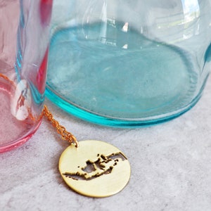 May include: A gold necklace with a pendant shaped like the outline of Indonesia. The pendant is hanging from a chain and is resting on a gray surface.