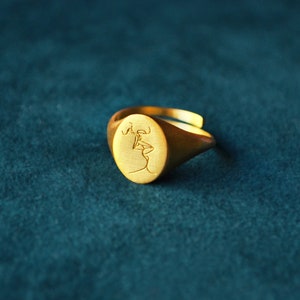 Puede incluir: Un anillo de sello de oro con un dise&ntilde;o grabado de dos figuras bes&aacute;ndose.