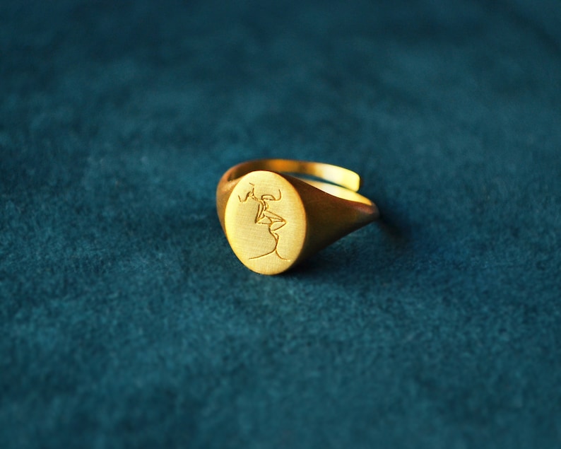 Puede incluir: Un anillo de sello de oro con un dise&ntilde;o grabado de dos figuras bes&aacute;ndose.