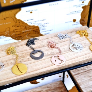 May include: Six necklaces with pendants shaped like different countries. The pendants are made of silver, gold, and black metal. The necklaces are displayed on a wooden shelf.