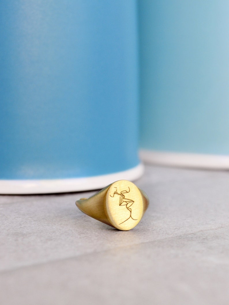 Puede incluir: Un anillo de sello ovalado de oro con un dibujo de l&iacute;nea de dos personas bes&aacute;ndose grabado en la cara.