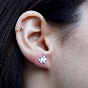 May include: A close-up of a person's ear with a small silver puzzle piece earring. The earring is a simple design and is a subtle way to add a touch of personality to any outfit.
