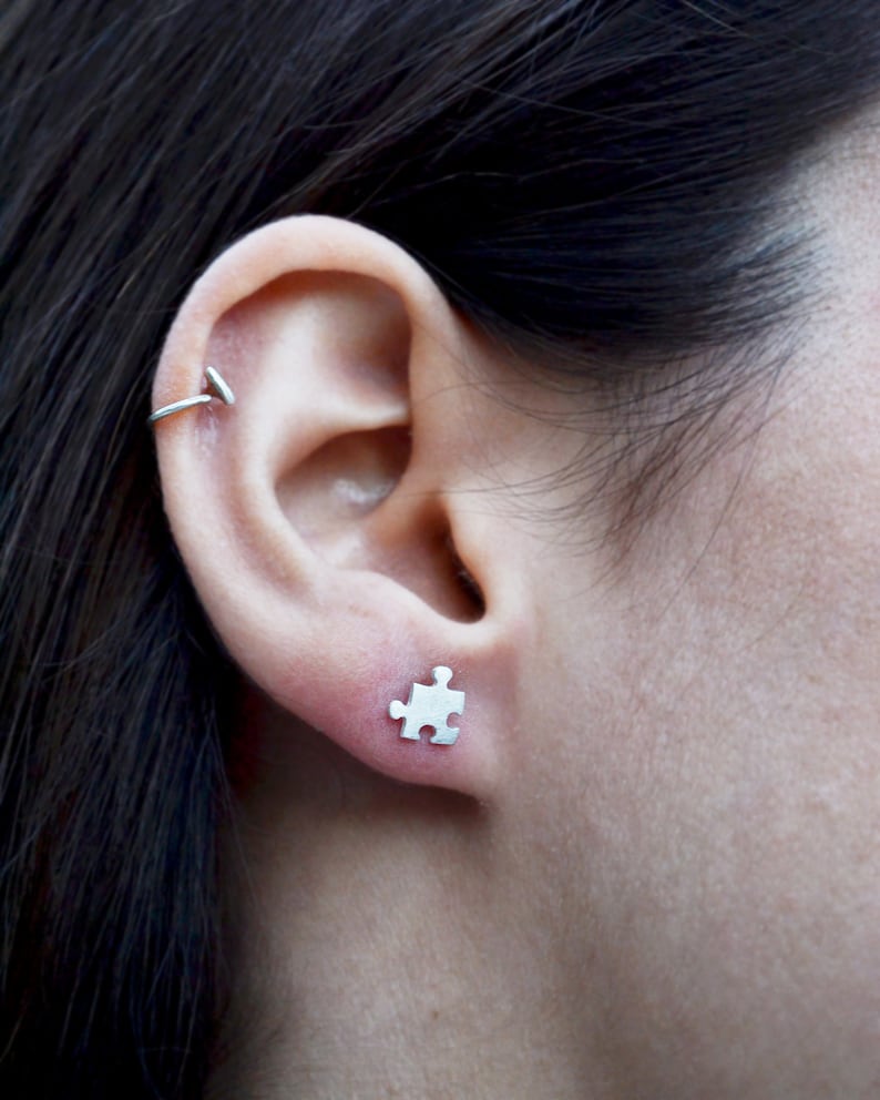 May include: A close-up of a person's ear with a small silver puzzle piece earring. The earring is a simple design and is a subtle way to add a touch of personality to any outfit.