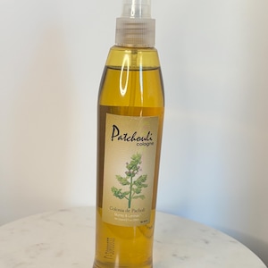 Puede incluir: Una botella de colonia de vidrio transparente llena de líquido ámbar. La etiqueta dice "Patchouli cologne" con una ilustración botánica de una planta. La parte inferior de la etiqueta dice "Colonia de Pacholi Murray & Lanman". La botella tiene un pulverizador.