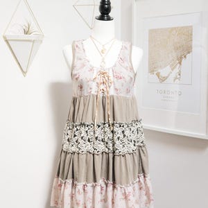 May include: A sleeveless dress on a mannequin. The dress features a floral print bodice, a lace midsection, and a tiered skirt. The colors are muted, with shades of pink, beige, and brown. A gold necklace is visible.