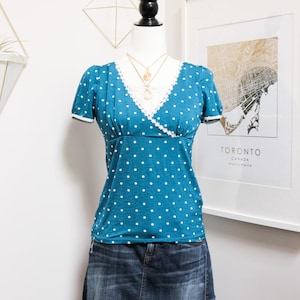 Peut inclure: Un haut turquoise à pois blancs avec un col en V et une bordure en dentelle blanche, associé à une jupe en jean. Le haut a des manches courtes et une taille ajustée. Un collier en or est visible. La tenue est présentée sur un mannequin.