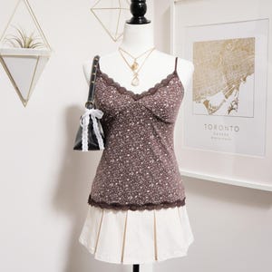 May include: A mannequin showcases a brown floral camisole with lace detailing, worn with a cream pleated skirt. A black handbag with a white bow and gold necklaces accessorise the outfit. A framed map of Toronto is visible in the background.