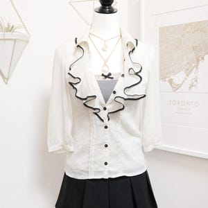 May include: A white blouse with black polka dots and a ruffled collar, paired with a black skirt. The blouse has black buttons and a black trim on the ruffles. A necklace with a pendant is visible.