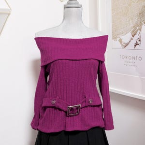 May include: A magenta off-the-shoulder sweater with long sleeves and a ribbed texture. The sweater features a silver buckle belt and is paired with a black pleated skirt. The sweater is displayed on a mannequin.