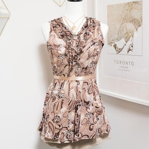 May include: A sleeveless paisley print top in shades of brown, beige, and white, with a ruffled neckline and a matching ribbon belt. The top is displayed on a mannequin with layered necklaces.