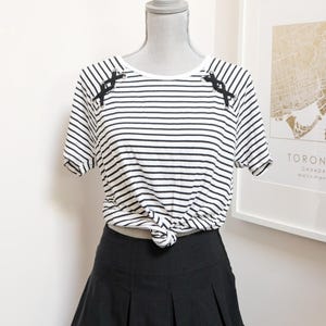 May include: A black and white striped t-shirt with black accents on the shoulders is knotted at the waist. Paired with a black pleated mini skirt, the outfit is displayed on a mannequin. The background includes a framed map.