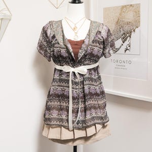 May include: A patterned, short-sleeved top with a white ribbon belt, layered over a brown camisole and a beige skirt. The top features a floral and geometric design in shades of purple, black, and beige. A gold necklace is visible.