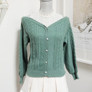 May include: A sage green cardigan with a cable knit design and pearl buttons. The cardigan is paired with a white lace top and a white pleated skirt. The outfit is displayed on a mannequin.