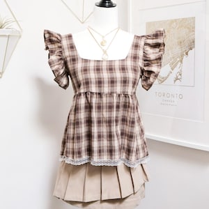 May include: A brown plaid top with frilled sleeves and a square neckline is displayed on a mannequin. The top is paired with a beige pleated skirt and a delicate lace trim. A gold necklace hangs on the mannequin.