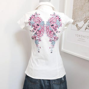 May include: White short-sleeved shirt with a floral butterfly design on the back in pink, blue, and purple. The shirt is paired with a denim skirt. The shirt is displayed on a mannequin.