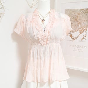 May include: A light pink blouse with short sleeves and a ruffled neckline is displayed on a mannequin. The blouse has a gathered waist and is paired with a white skirt. A gold necklace with a pendant is visible.