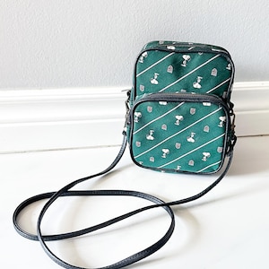 May include: Green crossbody bag with a repeating pattern of Snoopy and diagonal white stripes. The bag has a black strap and trim, with a zippered main compartment and a smaller front pocket. The bag is made of a durable fabric.
