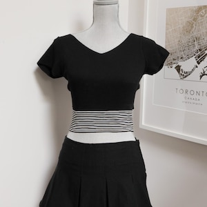 May include: A black two-piece outfit displayed on a mannequin. The top is a short-sleeved, V-neck design. The waistline features a horizontal striped band. The skirt is a pleated mini skirt. The outfit is all black.