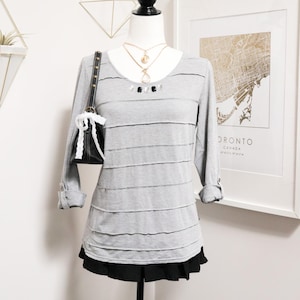 May include: A gray long-sleeve shirt with horizontal layered detailing, displayed on a mannequin. The shirt has a scoop neck and is paired with a black skirt. A black purse with white accents hangs on the mannequin's arm, and necklaces adorn the neck.