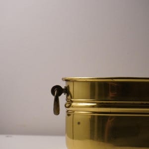 Puede incluir: Un recipiente de latón antiguo con un asa. El recipiente tiene forma rectangular con esquinas redondeadas y un acabado brillante y pulido. El asa está sujeta al lateral y es de un metal más oscuro. Es probable que el recipiente se utilice para guardar cosas.