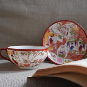 Op de afbeelding: Een vintage theekop en schotel set met een Japans geïnspireerd ontwerp. De kop en schotel hebben een rode rand en gedetailleerde illustraties van figuren en landschappen. Een open boek staat op de voorgrond.