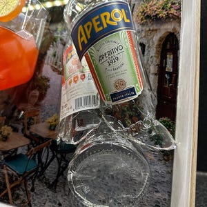 May include: Aperol bottle with a label featuring the text "APERITIVO DAL 1919" and "PADOVA-ITALIA". The bottle is partially broken, with a background of a street scene and a glass of orange-colored liquid with an orange slice.
