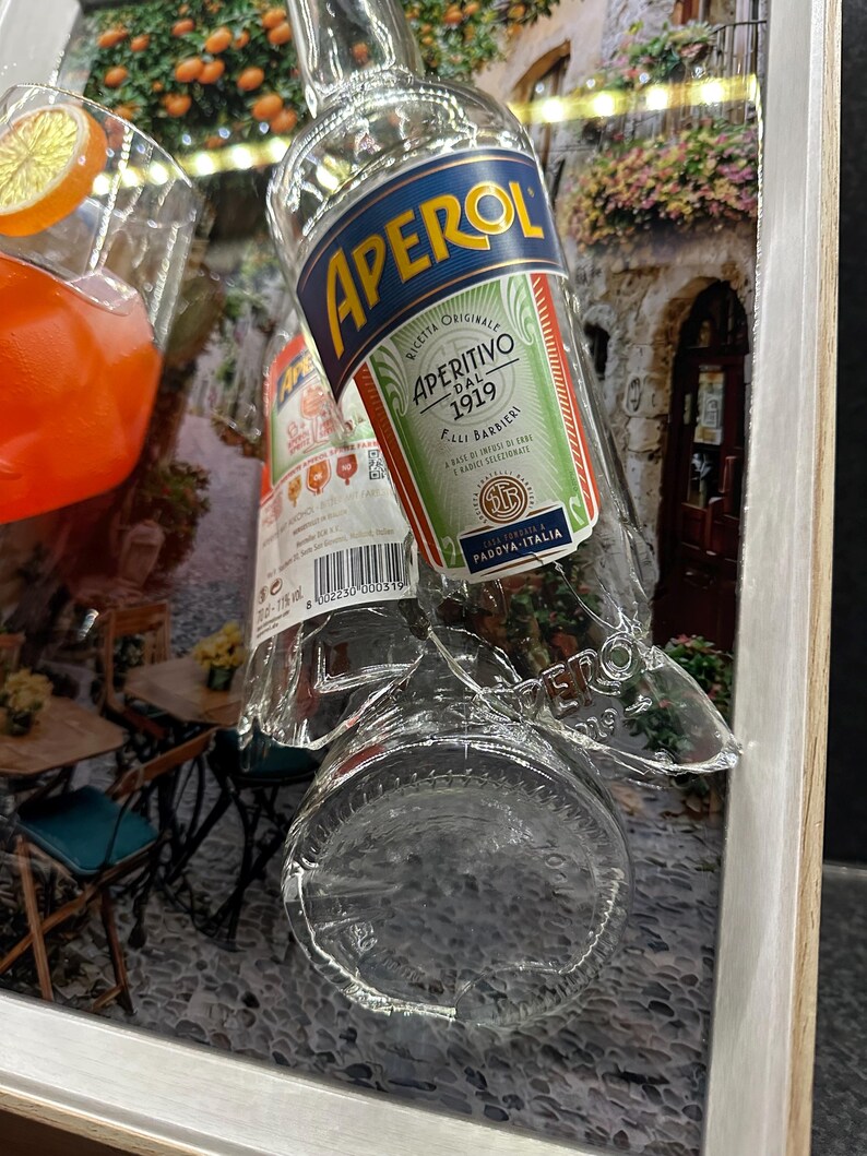 May include: Aperol bottle with a label featuring the text "APERITIVO DAL 1919" and "PADOVA-ITALIA". The bottle is partially broken, with a background of a street scene and a glass of orange-colored liquid with an orange slice.