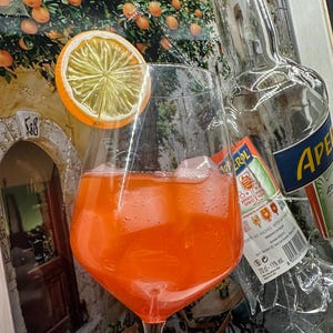 May include: Aperol Spritz cocktail in a large wine glass with ice and an orange slice. A partially visible Aperol bottle is behind the glass. The background features a painting of an orange grove and a stone building.
