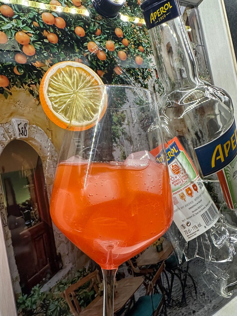 May include: Aperol Spritz cocktail in a large wine glass with ice and an orange slice. A partially visible Aperol bottle is behind the glass. The background features a painting of an orange grove and a stone building.
