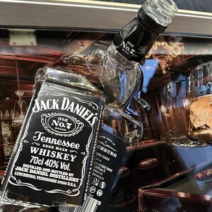 May include: A close-up of a Jack Daniel's Tennessee Whiskey bottle with the text "Old No. 7 Brand" and "70cl 40% Vol." The bottle is black and white with a clear glass top. The background includes a pool table and bar setting.