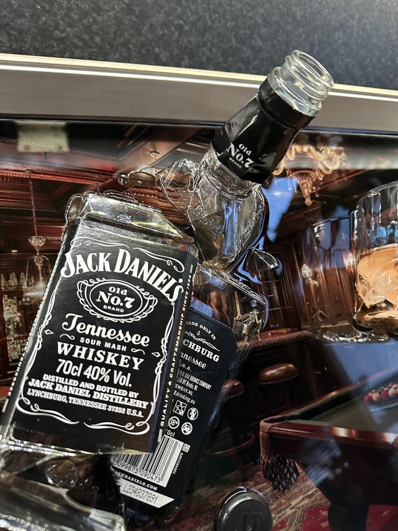 May include: A close-up of a Jack Daniel's Tennessee Whiskey bottle with the text "Old No. 7 Brand" and "70cl 40% Vol." The bottle is black and white with a clear glass top. The background includes a pool table and bar setting.