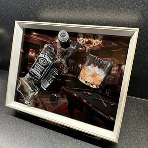 Op de afbeelding: Een ingelijst kunstwerk met een barscène met een Jack Daniel's fles, een glas whisky met ijs en een pooltafel. Het kunstwerk heeft een witte lijst en toont een zwak verlichte kamer met een kroonluchter.