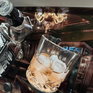 May include: A clear glass filled with ice cubes and a light orange liquid, next to a partially visible black bottle. The background shows a reflection of a room with a chandelier and pool table.