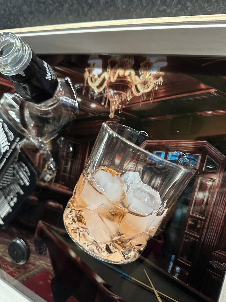 May include: A clear glass filled with ice cubes and a light orange liquid, next to a partially visible black bottle. The background shows a reflection of a room with a chandelier and pool table.