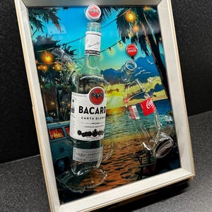 Op de afbeelding: Ingelijst kunstwerk met een strandscène met een Bacardi Carta Blanca-fles en een Coca-Cola-fles. De flessen zijn gedeeltelijk gebroken, met doppen en een tropische achtergrond. De lijst is licht van kleur.