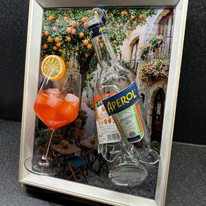Op de afbeelding: Een ingelijst kunstwerk met een glas sinaasappelcocktail met ijs en een schijfje sinaasappel, naast een gedeeltelijk zichtbare Aperol-fles. De achtergrond toont een Europese straatscène met sinaasappelbomen en een café.