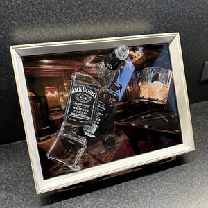 May include: A framed artwork featuring a Jack Daniel's whiskey bottle, a glass of whiskey with ice, and a pool table scene. The artwork has a wooden frame and a glass front, creating a 3D effect. The bottle is partially broken.