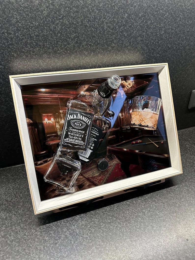 May include: A framed artwork featuring a Jack Daniel's whiskey bottle, a glass of whiskey with ice, and a pool table scene. The artwork has a wooden frame and a glass front, creating a 3D effect. The bottle is partially broken.