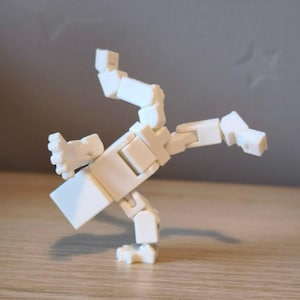 Pode incluir: Uma figura branca articulada numa pose dinâmica, parecendo estar em ação. A figura é feita de peças retangulares e cúbicas conectadas, com juntas visíveis. A figura está sobre uma superfície de madeira.
