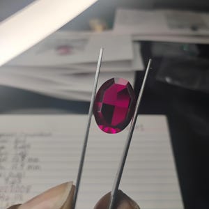 May include: A faceted, oval-shaped, deep red gemstone is held by tweezers. The gem is a vibrant magenta color, reflecting light. The background includes a hand, a notepad with handwritten notes, and a bright light.