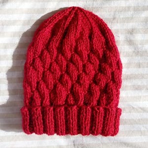 May include: A vibrant red knitted beanie featuring a textured cable knit pattern throughout the body and a thick, ribbed cuff at the base. The hat is constructed from yarn with a consistent, saturated red colour and a structured, rounded top shape.
