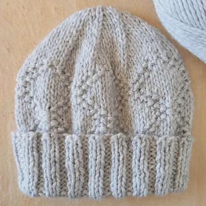May include: A light grey knitted beanie featuring a ribbed cuff and a textured pattern of vertical columns with small, raised knit stitches. The hat is crafted from thick yarn and displays a consistent, neutral colour throughout the entire design.