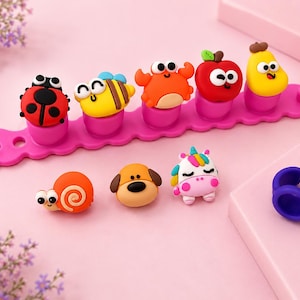 May include: A bright pink silicone bracelet band featuring five interchangeable charms shaped like a ladybug, bee, crab, apple, and pear. Below are three loose charms shaped like a snail, dog, and unicorn, alongside a single purple silicone ring.