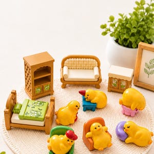 Peut inclure: Un ensemble de meubles de maison de poupée miniature comprenant un lit en bois avec une couverture verte à motifs, une grande armoire, un canapé et une petite table. Six figurines de canetons jaunes sont disposées autour des meubles.
